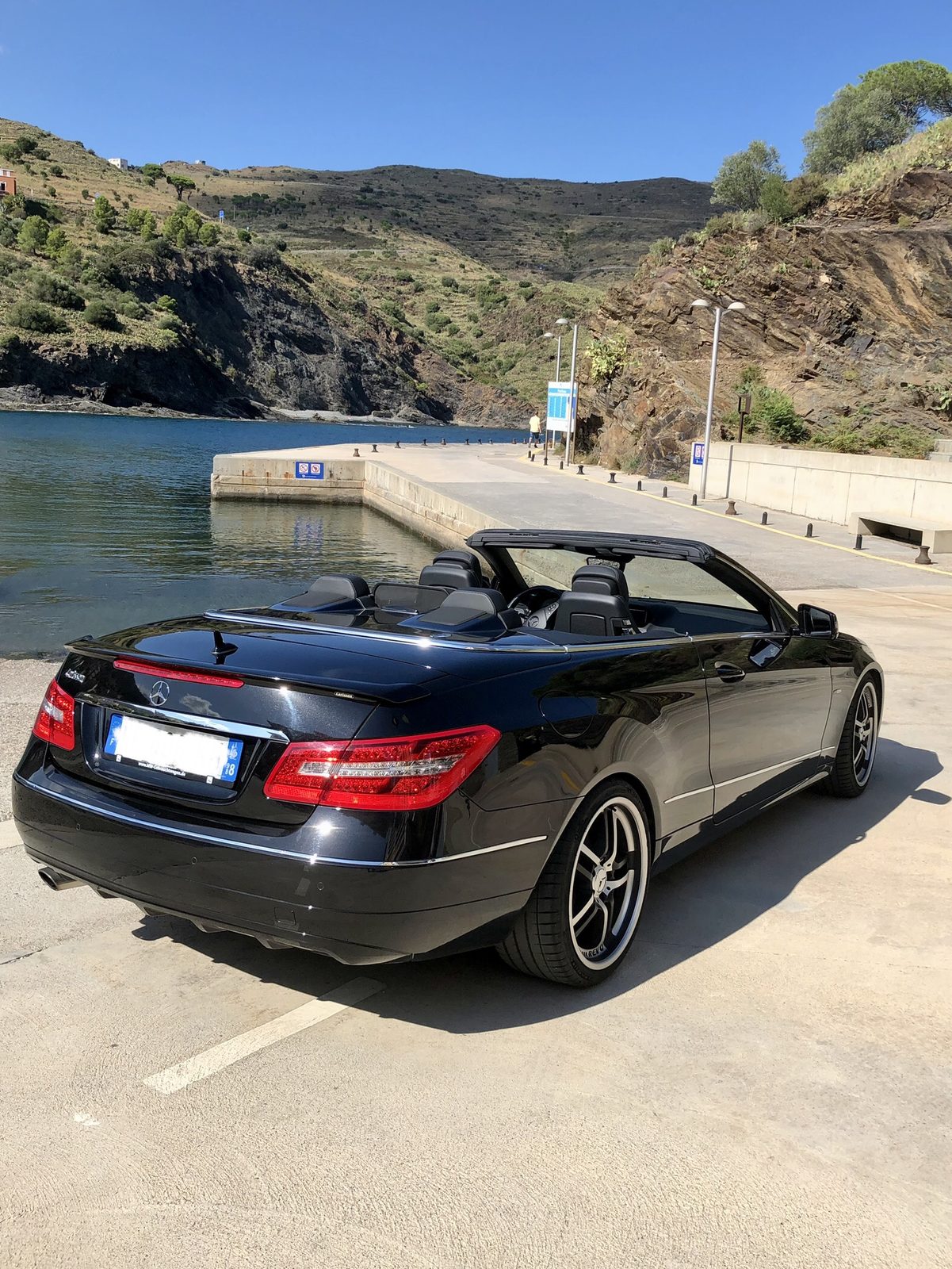 La Mercedes E 250 Cabriolet Carlsson noire, capote ouverte, devant un port de la côte catalane